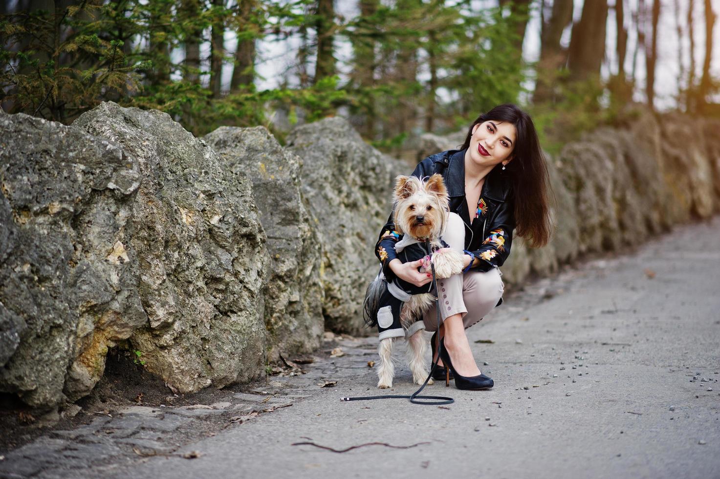 brunett zigenare tjej med yorkshire terrier hund poserade mot stenar på park. modell slitage på skinnjacka och t-shirt med ornament, byxor och skor med höga klackar. foto