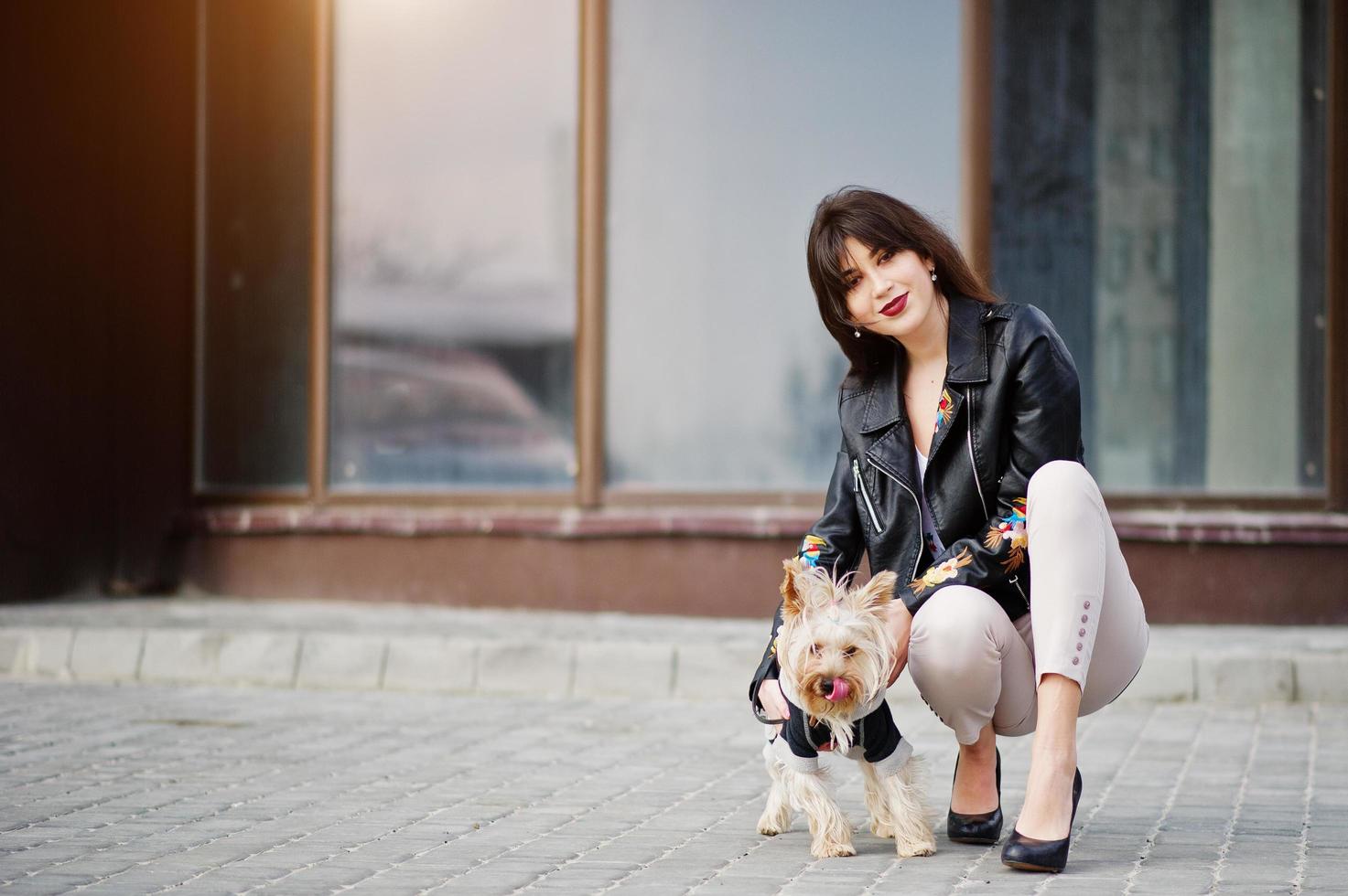 brunett zigenare tjej med yorkshire terrier hund poserade mot stora fönster hus. modell slitage på skinnjacka och t-shirt med ornament, byxor och skor med höga klackar. foto