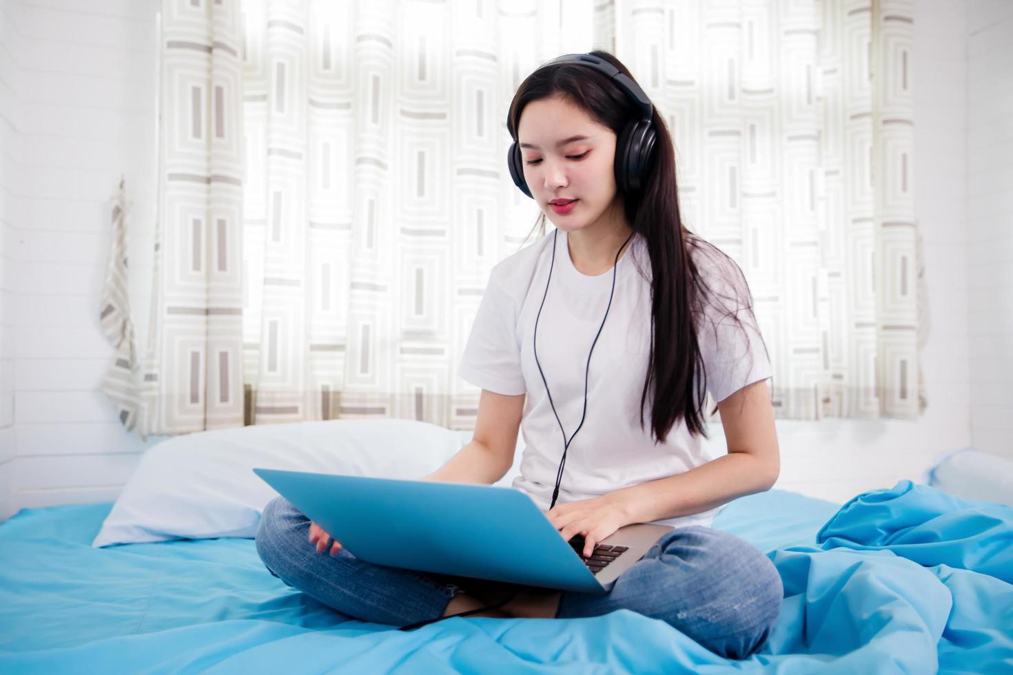 asiatisk glad ung kvinna bär hörlurar och lyssnar på musik sång och tittar på webbplats för att shoppa online från bärbar dator på sängen hemma foto