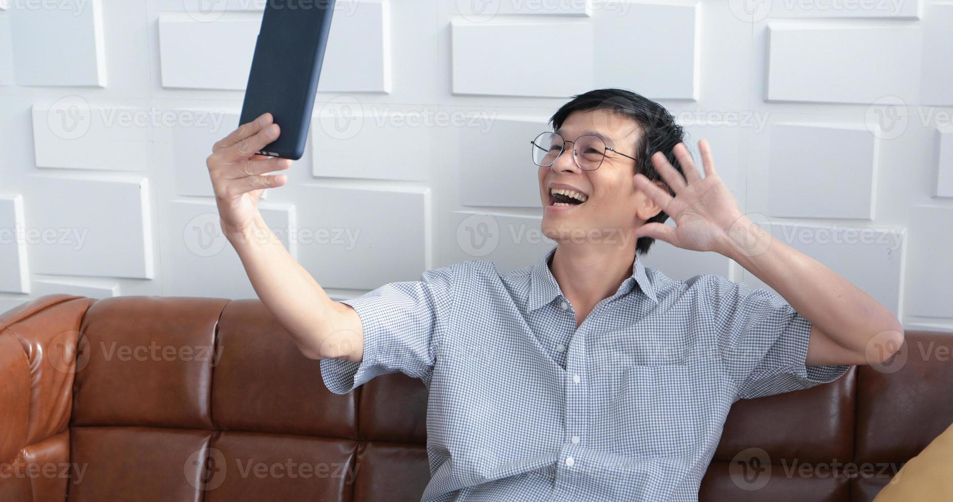 senior asiatisk man spelar surfplatta och videosamtal på soffan i vardagsrummet hemma porträtt av asiatisk äldre man är avkopplande och lycka med att spela surfplatta foto