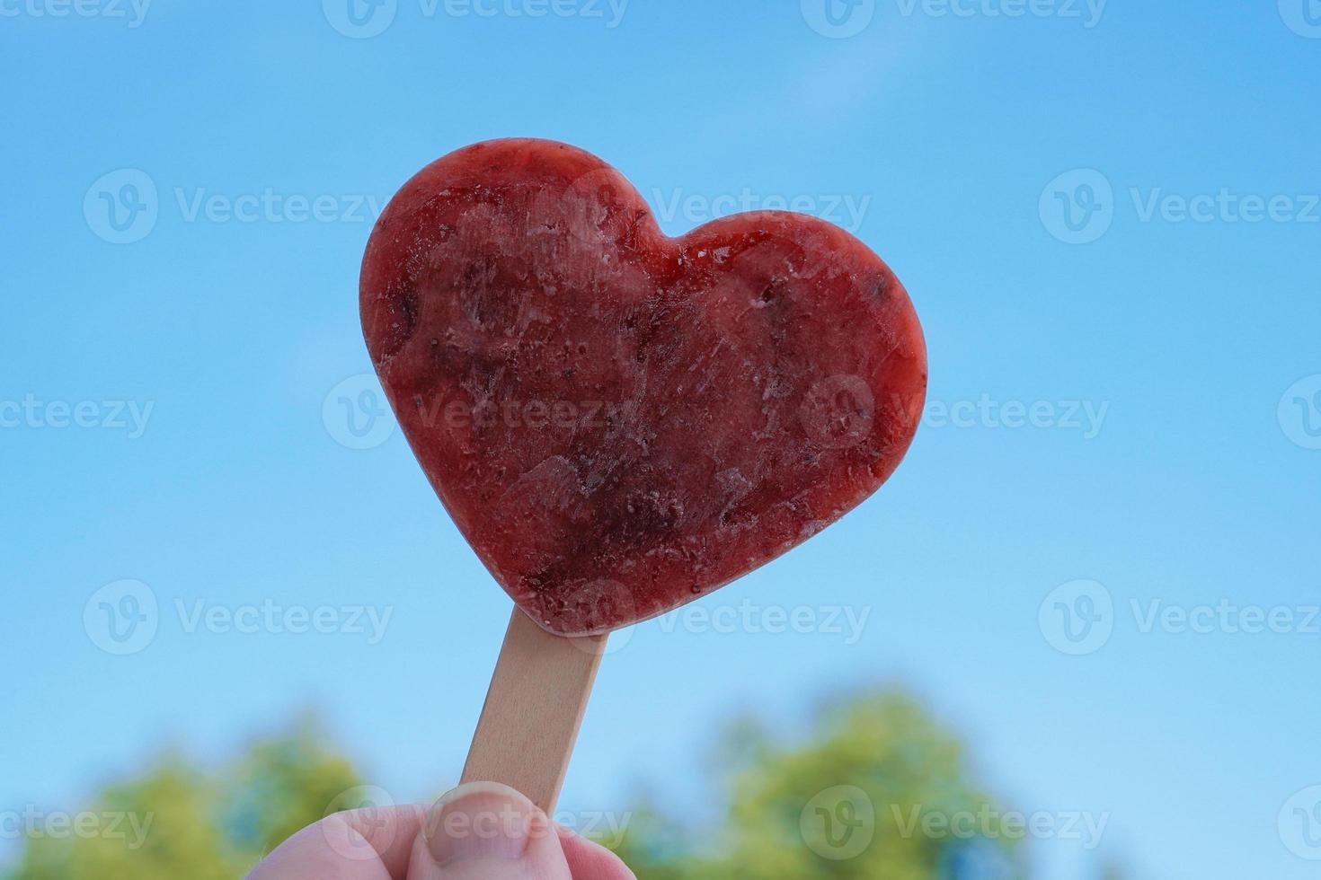 hand som håller hjärtformad popsicle eller ispinne mot klarblå himmel foto