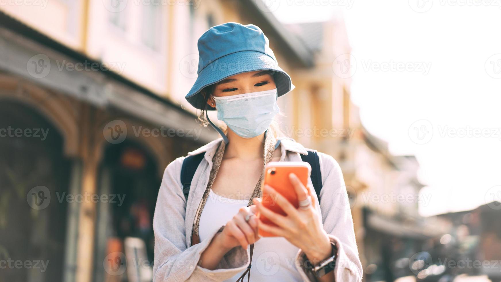 ung vuxen asiatisk kvinna ryggsäck resenären bära ansiktsmask och använda mobiltelefon för ansökan. foto