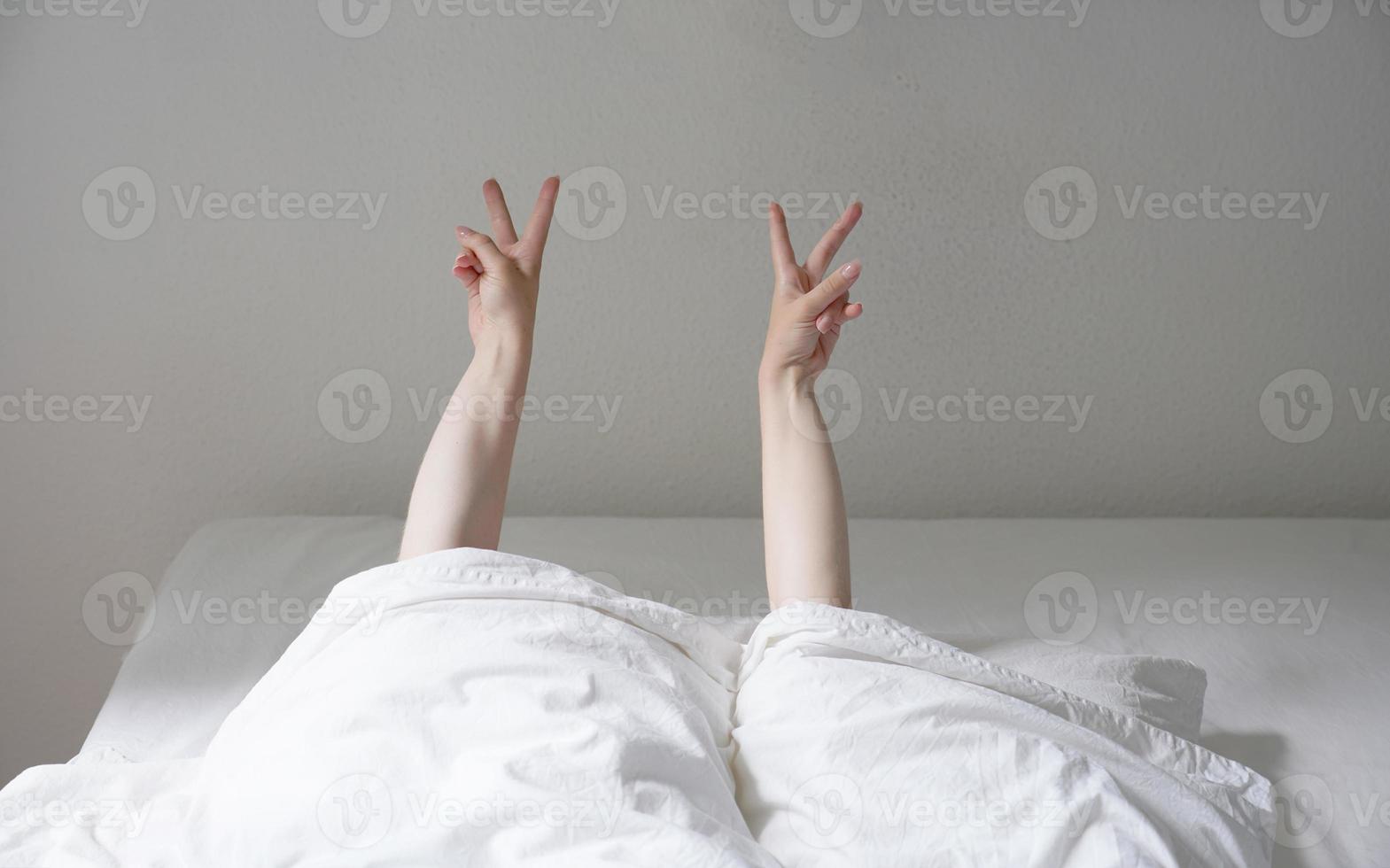 kvinna som visar fred eller v hand tecken medan liggande i mysig säng och gömmer sig under täcket foto