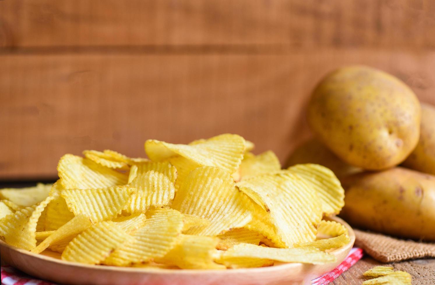 potatischips mellanmål på tallrik, krispiga potatischips på köksbordet och färsk rå potatis på träbakgrund foto