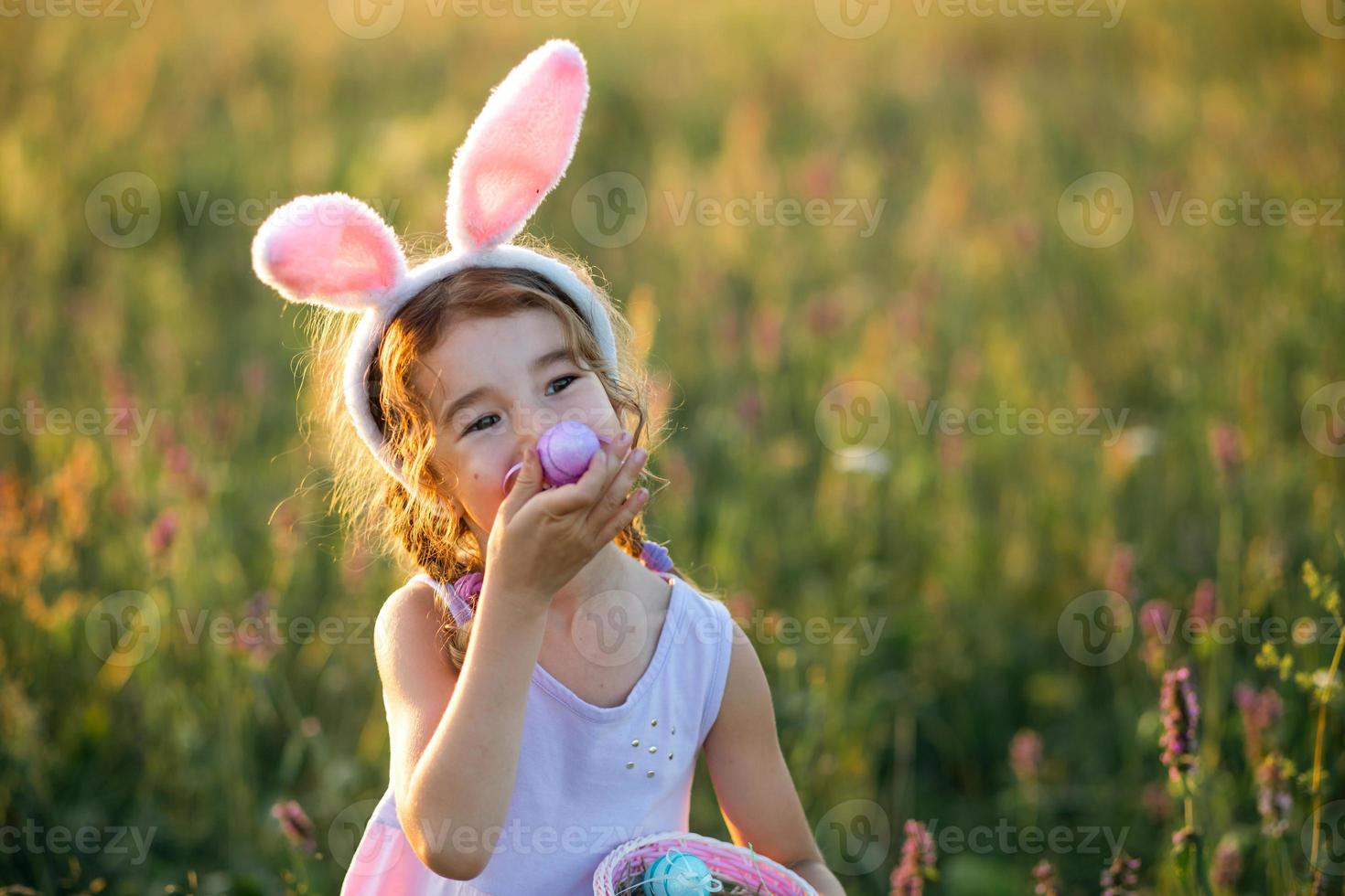 söt rolig tjej med målade påskägg på våren i naturen i ett fält med gyllene solljus och blommor. påskhelg, påskharen med öron, färgglada ägg i en korg. livsstil foto