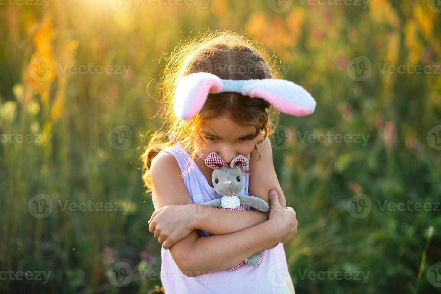 söt 5-årig tjej med kaninöron kramar försiktigt en leksakskanin i naturen i ett blommande fält på sommaren med gyllene solljus. påsk, påskhare, barndom, lyckligt barn, våren. foto