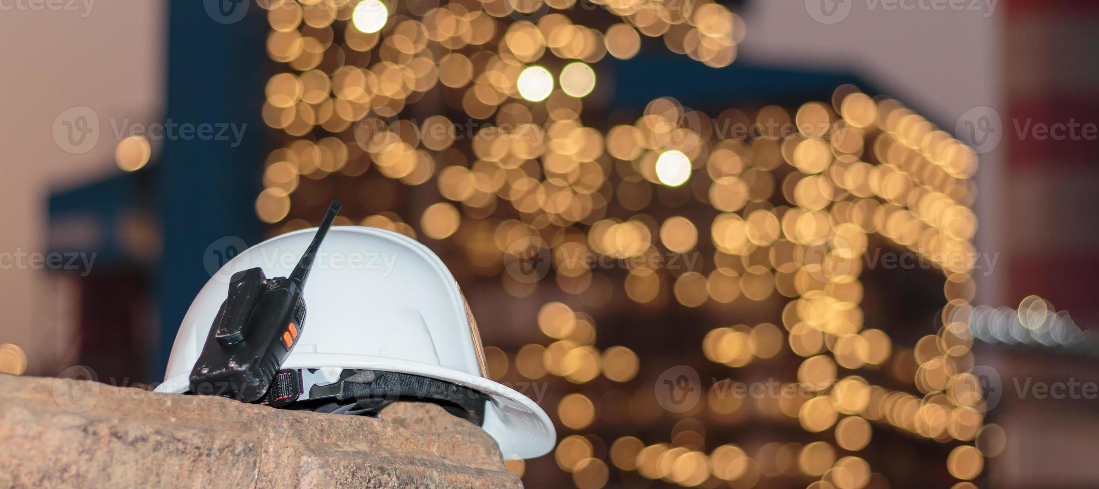 en vit hjälm och radio kommunicerar med en bokeh-bakgrund, tänder ett kraftverk eller en fabrik i skymningen. foto