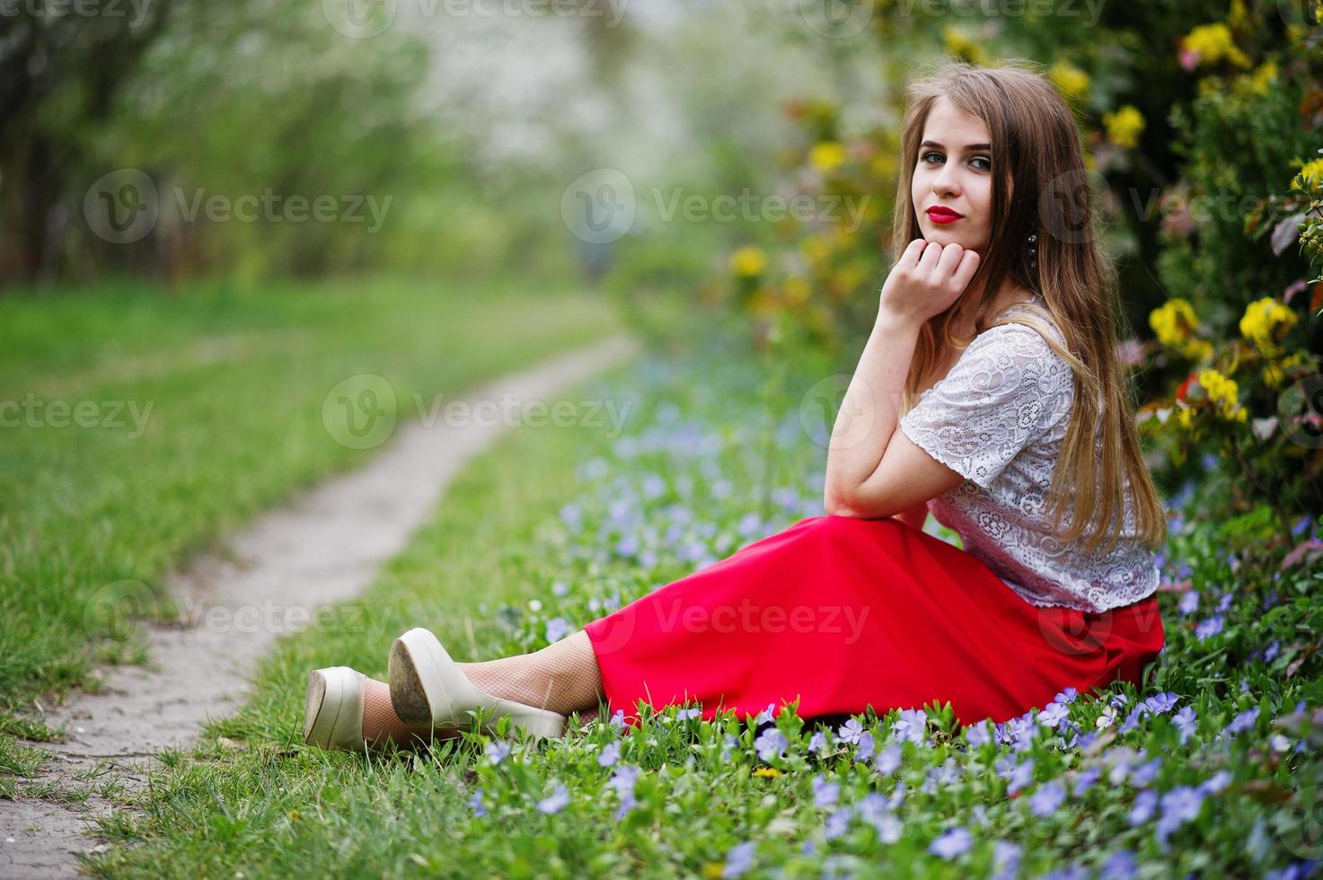 porträtt av sitiing vacker flicka med röda läppar på våren blossom trädgård på gräs med blommor, slitage på röd klänning och vit blus. foto