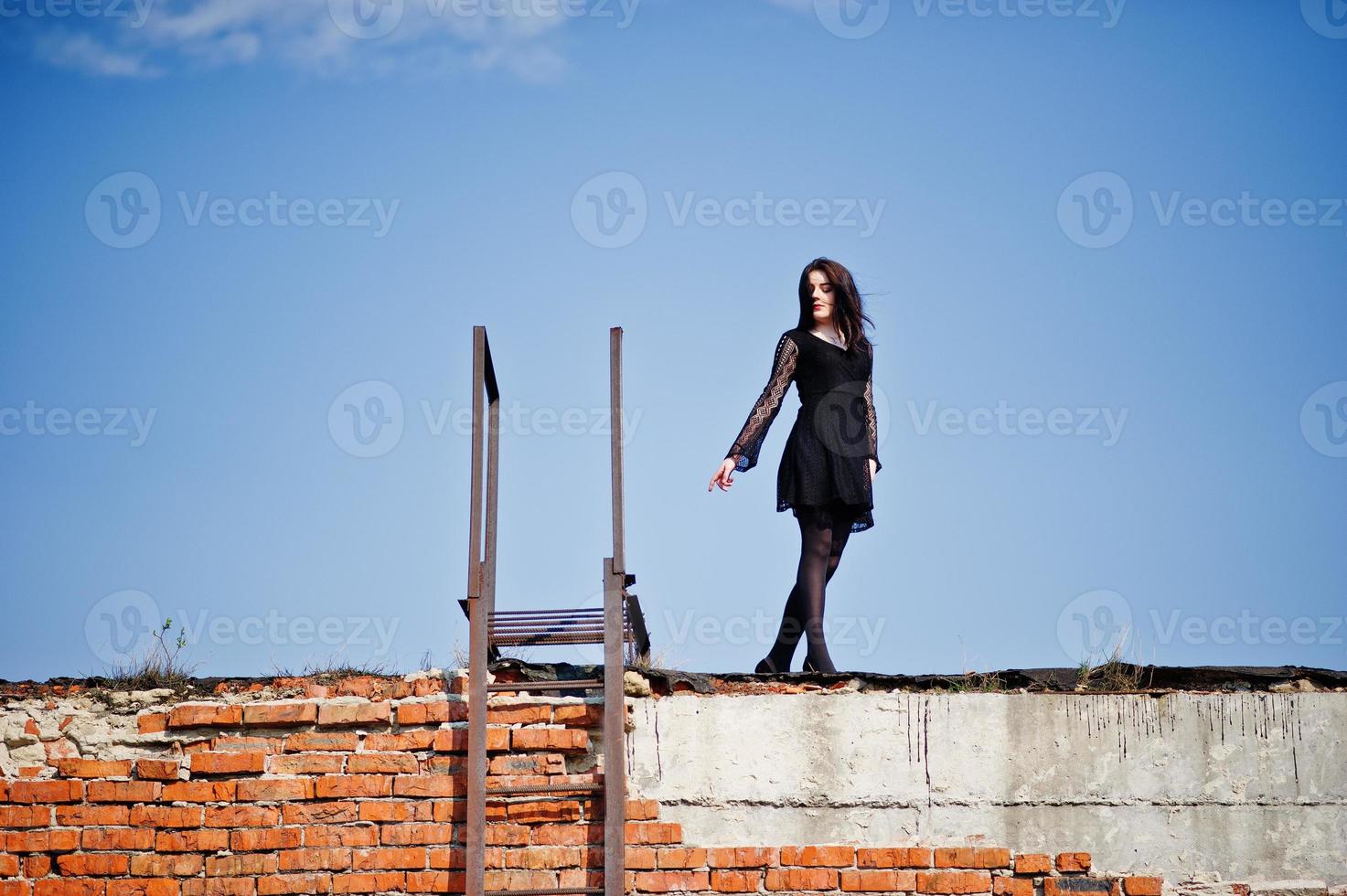 porträtt brunett tjej med röda läppar klädd i en svart klänning poserade på taket nära stegen. gatumodell. foto