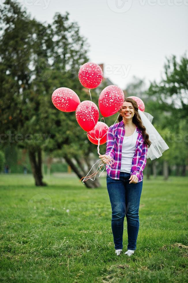 porträtt av brunett tjej på rutig skjorta, jeans och slöja med många röda ballonger på möhippo. foto