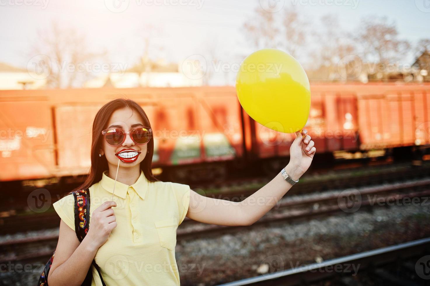 ung tonårsflicka står på perrongen vid tågstationen med roliga läppar på pinne och ballong till hands, bär på gul t-shirt, jeans och solglasögon, med ryggsäck. foto