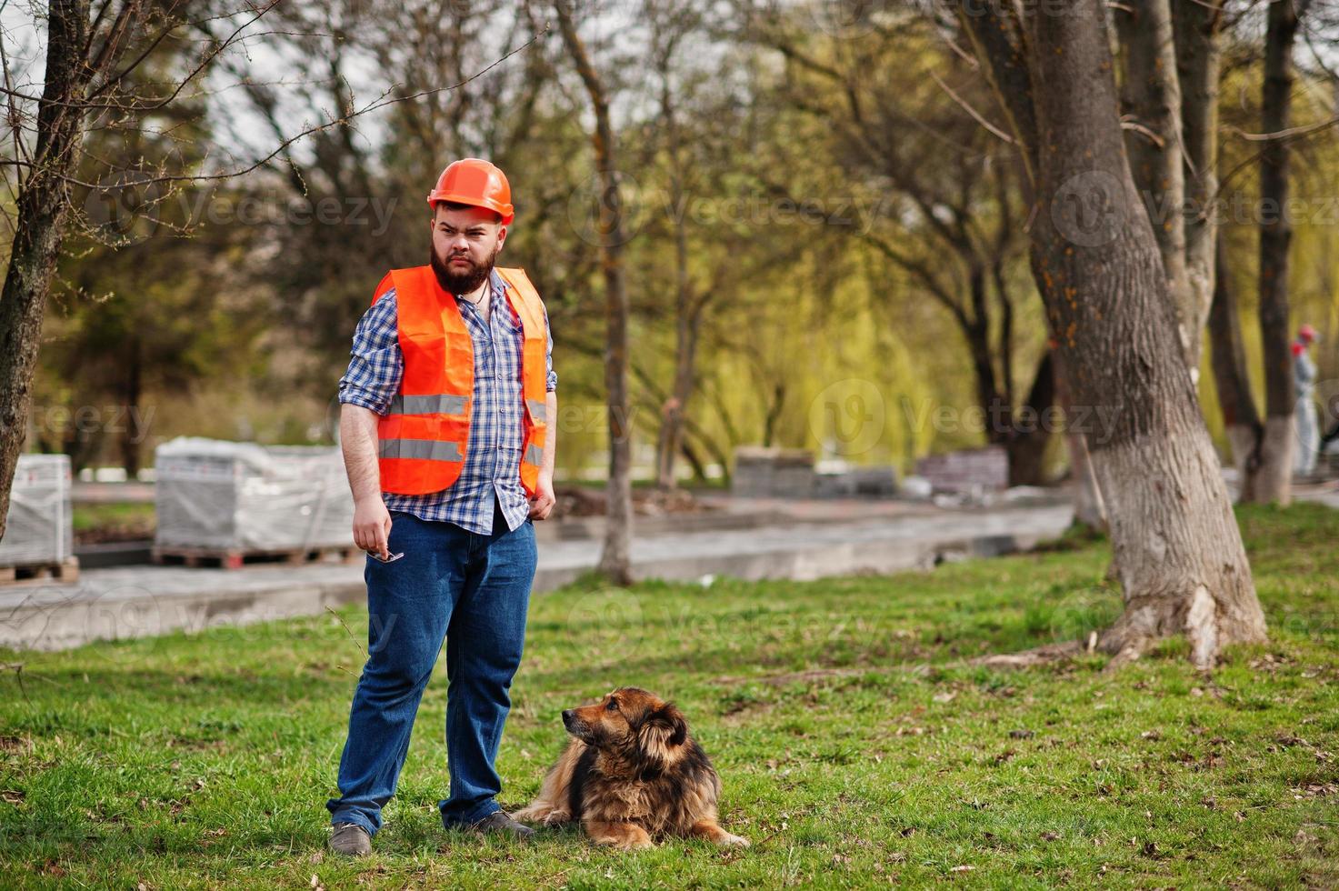 porträtt av brutala skägg arbetare man kostym byggnadsarbetare i säkerhet orange hjälm med vakthund. foto