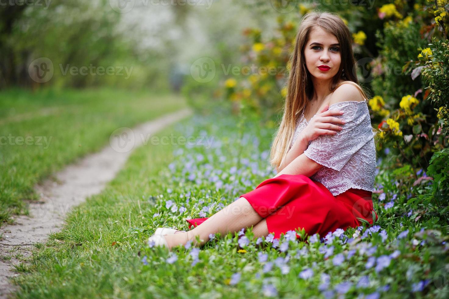 porträtt av sitiing vacker flicka med röda läppar på våren blossom trädgård på gräs med blommor, slitage på röd klänning och vit blus. foto