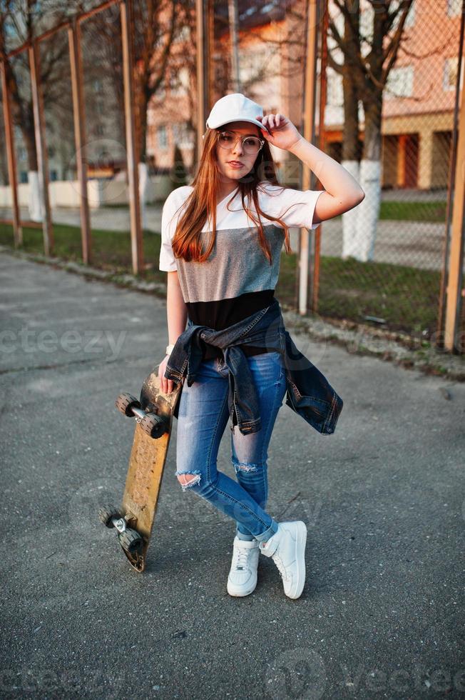 ung tonårig urban flicka med skateboard, slitage på glasögon, keps och trasiga jeans på gården idrottsplats. foto