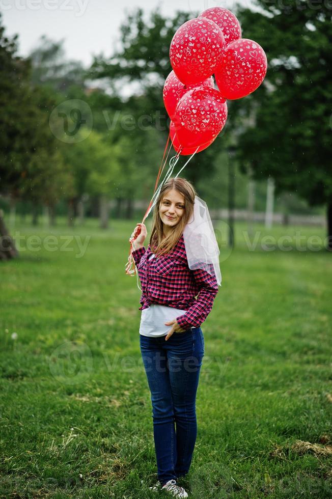 porträtt av brunett tjej på rutig skjorta, jeans och slöja med många röda ballonger på möhippo. foto