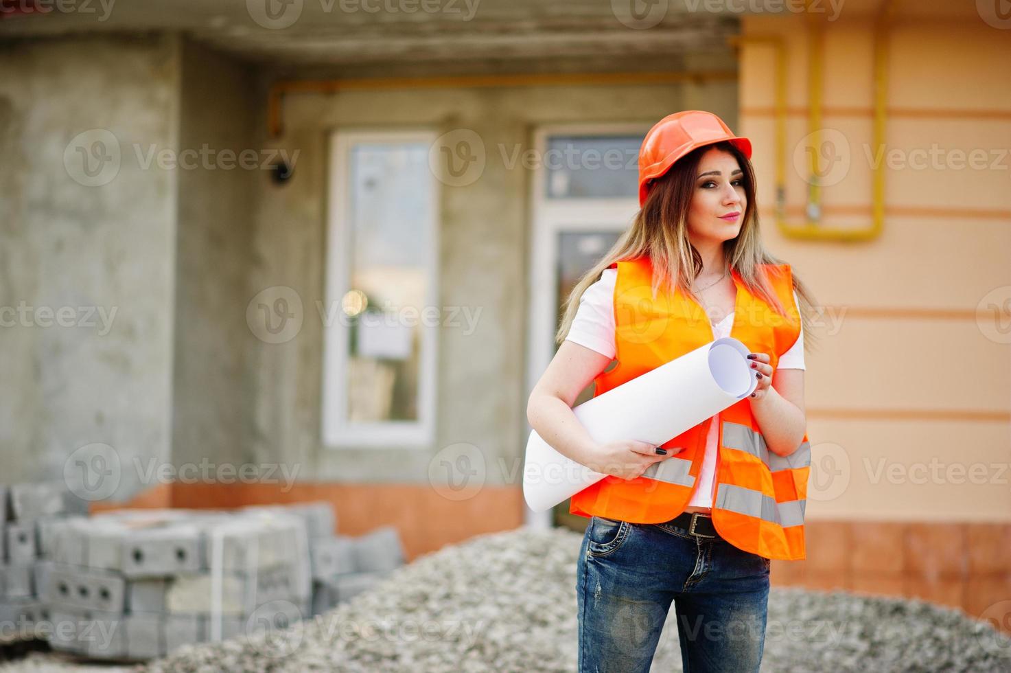 ingenjör byggare kvinna i uniform väst och orange skyddshjälm håller affärsritning pappersrulle mot ny byggnad. fastighet boende block tema. foto