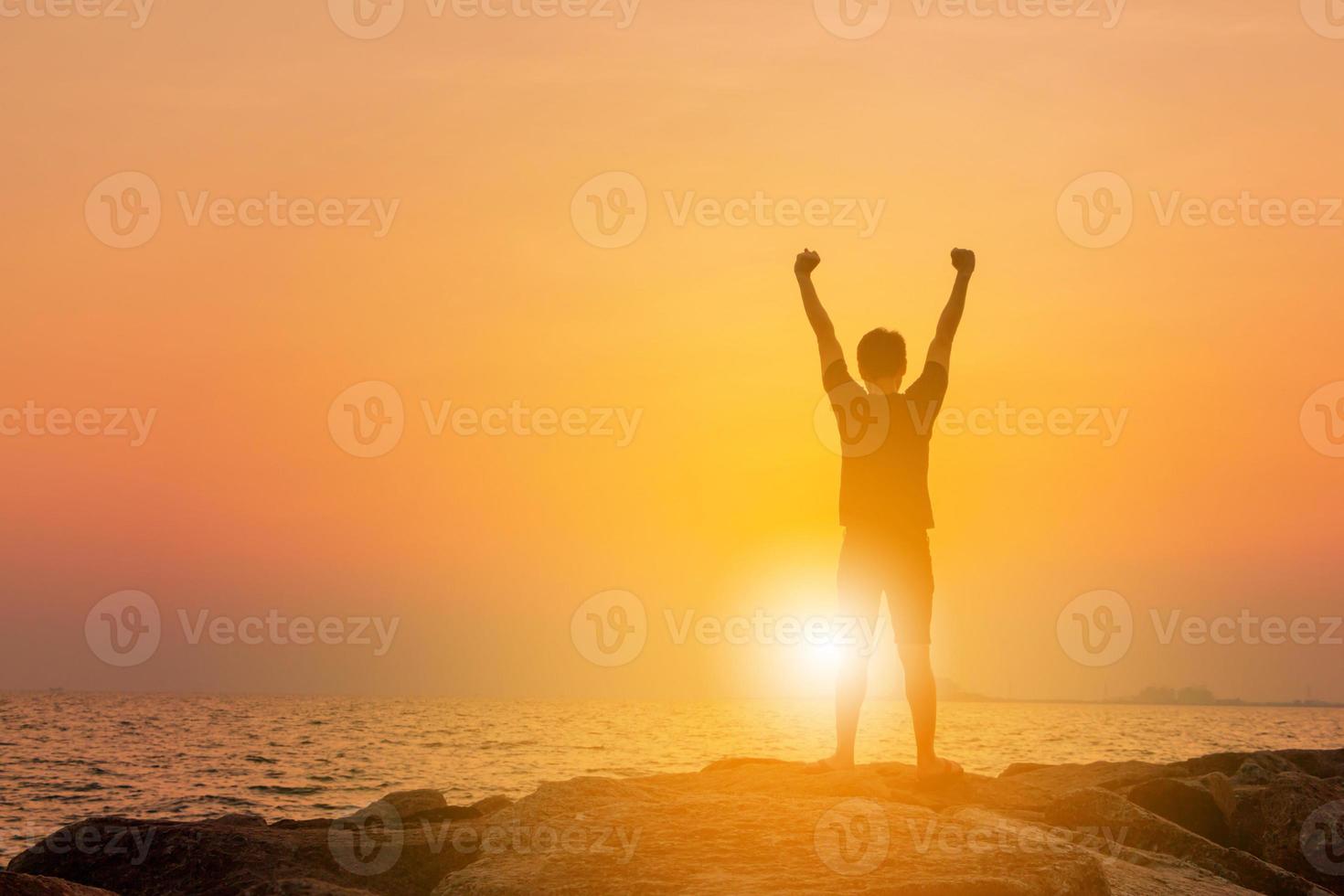 siluett av man som står med öppen hand på en sten och tittar på den gyllene solen. kvällshimmel solnedgång på stranden bakgrund, sport och njuta av livet på havet koncept. foto