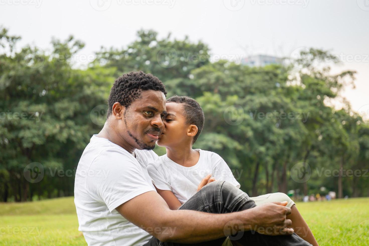 afrikansk söt liten pojke i casual kysser pappa i parken. glad afro-amerikansk far medan krama och bära sin son sitter utomhus, glad svart familj koncept. foto