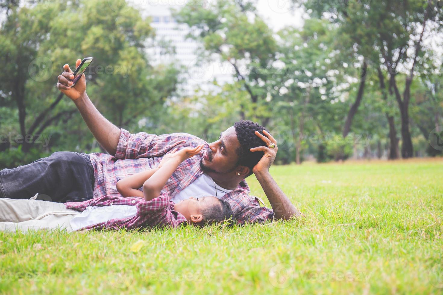 lycklig far med liten son som tar selfiebild med smartphone, glad afrikansk amerikansk far och söner som leker i parken foto