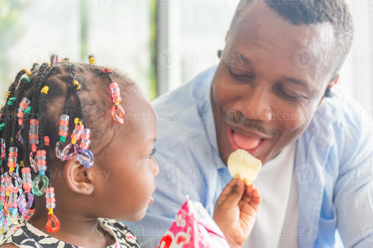 söt liten flicka tittar på hennes pappa som äter mellanmål, glada afroamerikanska far och dotter som leker i vardagsrummet, familjekoncept för lycka foto