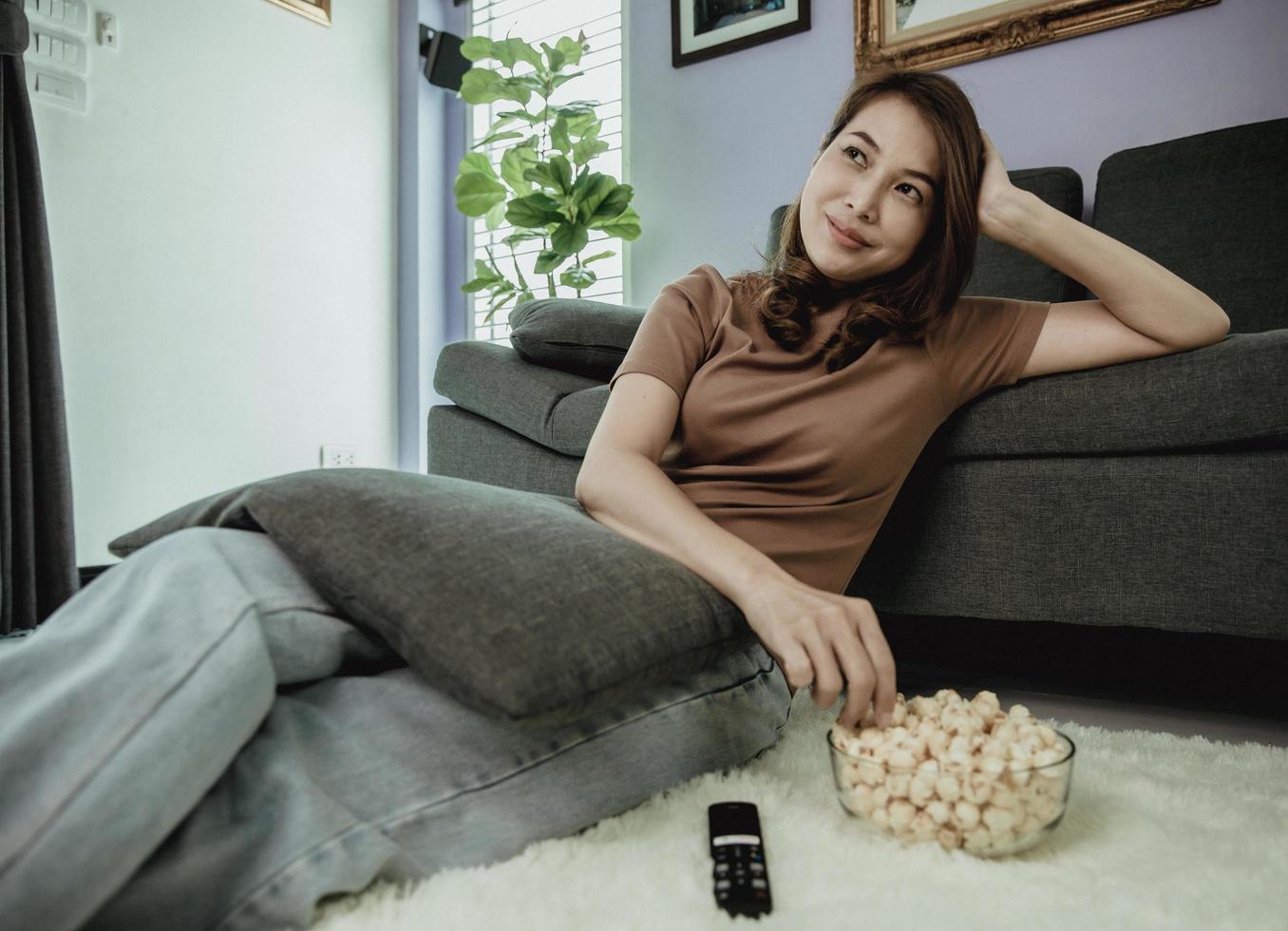 asiatisk vacker vuxen 45-talskvinna som sitter och lutar bäddsoffan i vardagsrummet och skål med popcorn och fjärrkontroll njuter av att titta på tv med glad foto
