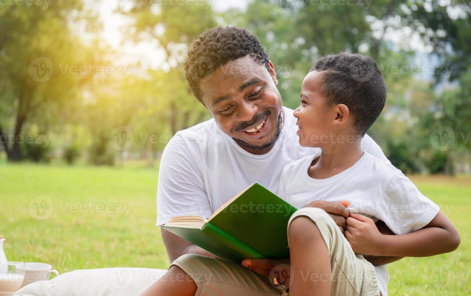 glad afrikansk amerikansk pojke och pappa som har en picknick i parken, glad son och pappa läser en bok, lycka familjekoncept foto