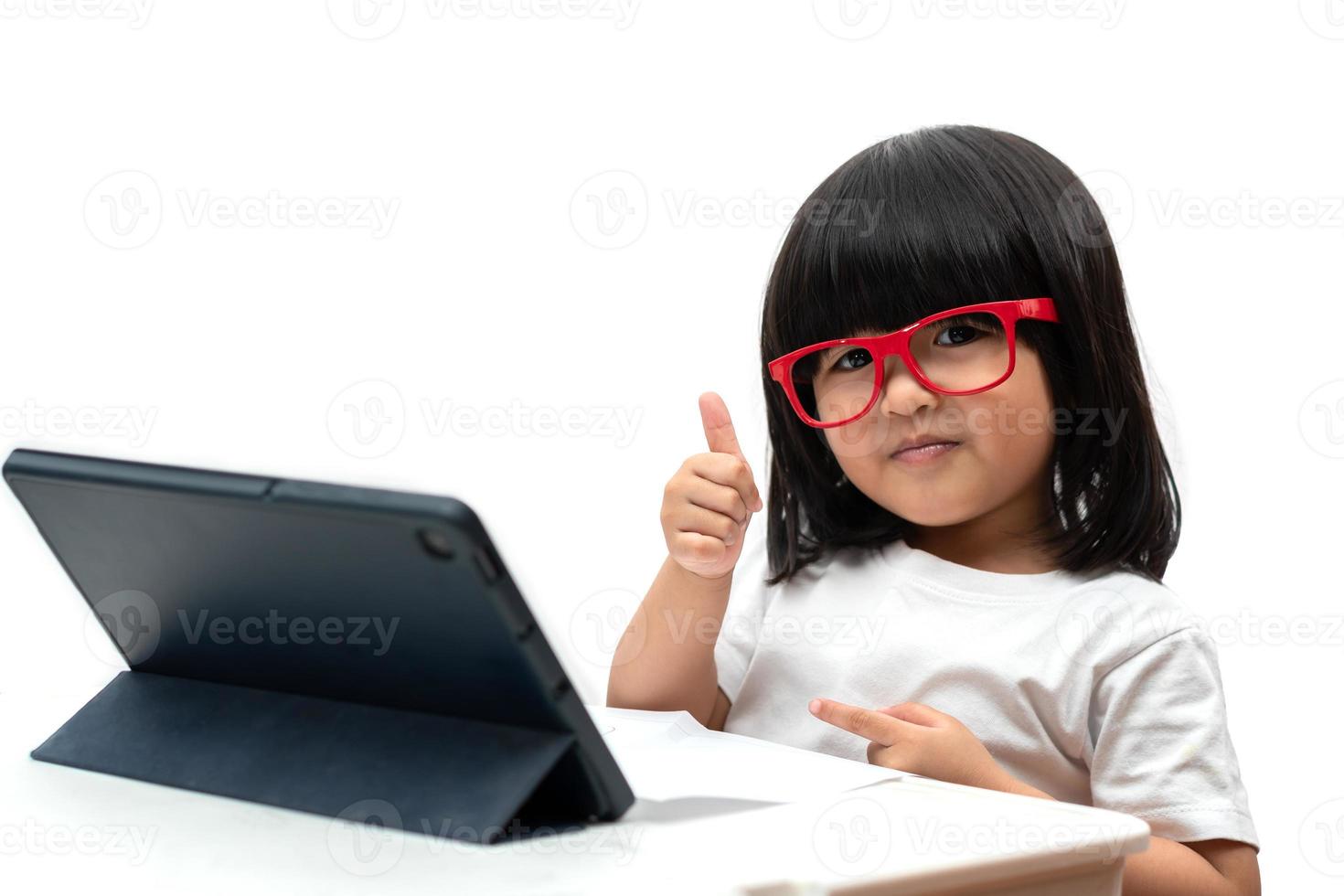 liten asiatisk förskolebarnflicka som bär röda glasögon och använder surfplatta på vit bakgrund och tummen upp, asiatisk tjej lär sig med ett videosamtal med surfplatta, pedagogiskt koncept för skolbarn foto