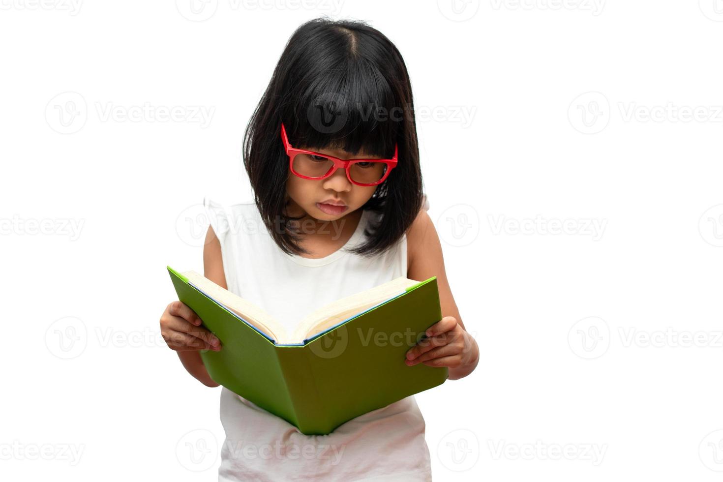 glad asiatisk liten förskoleflicka som bär röda glasögon håller och läser en grön bok på vit isolerad bakgrund. begreppet skolbarn och utbildning i grundskolan och förskolan, hemskola foto