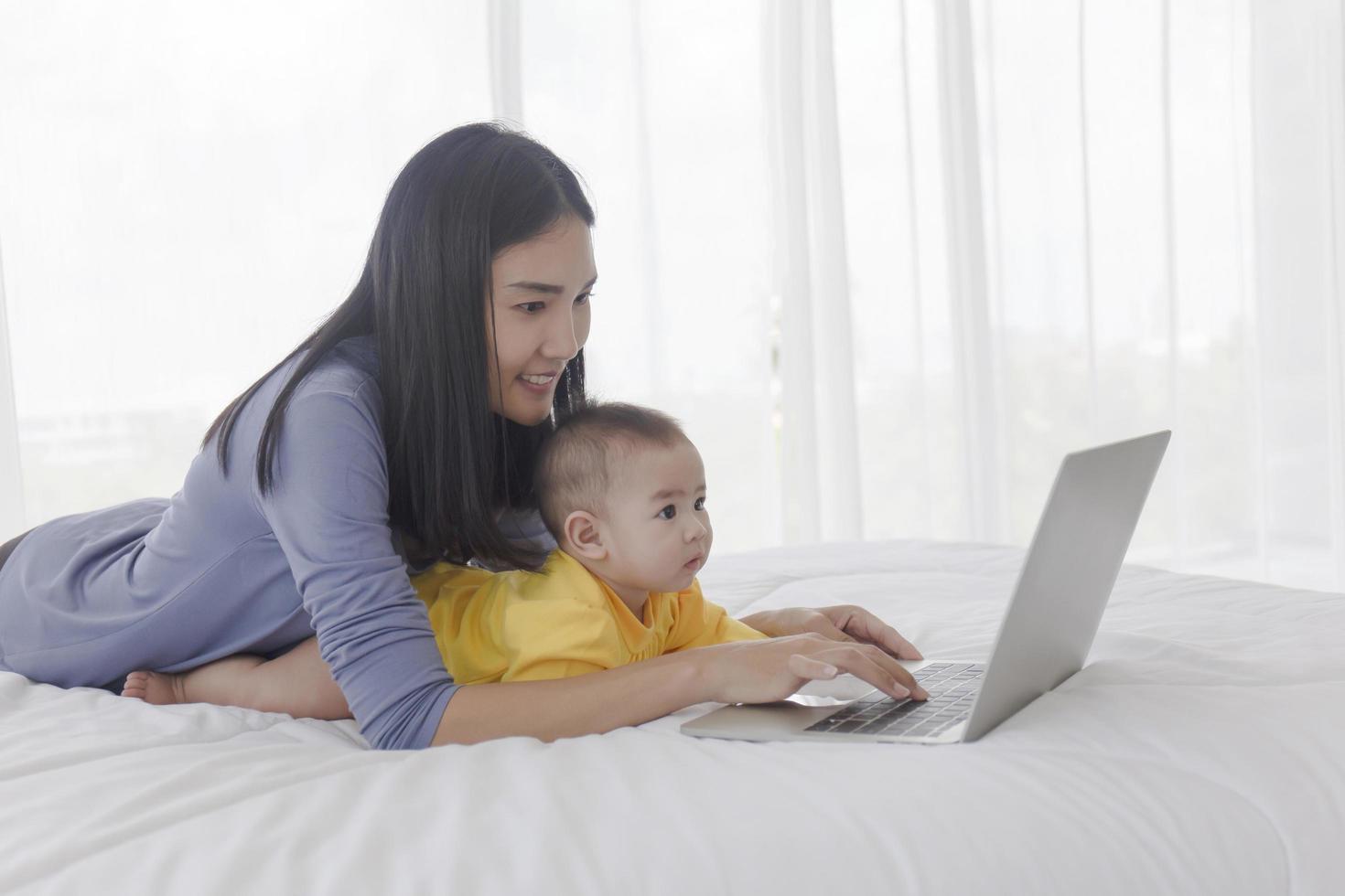 en asiatisk mamma arbetar på en bärbar dator i sängen och med sin bebis bredvid konceptet att arbeta hemma. foto