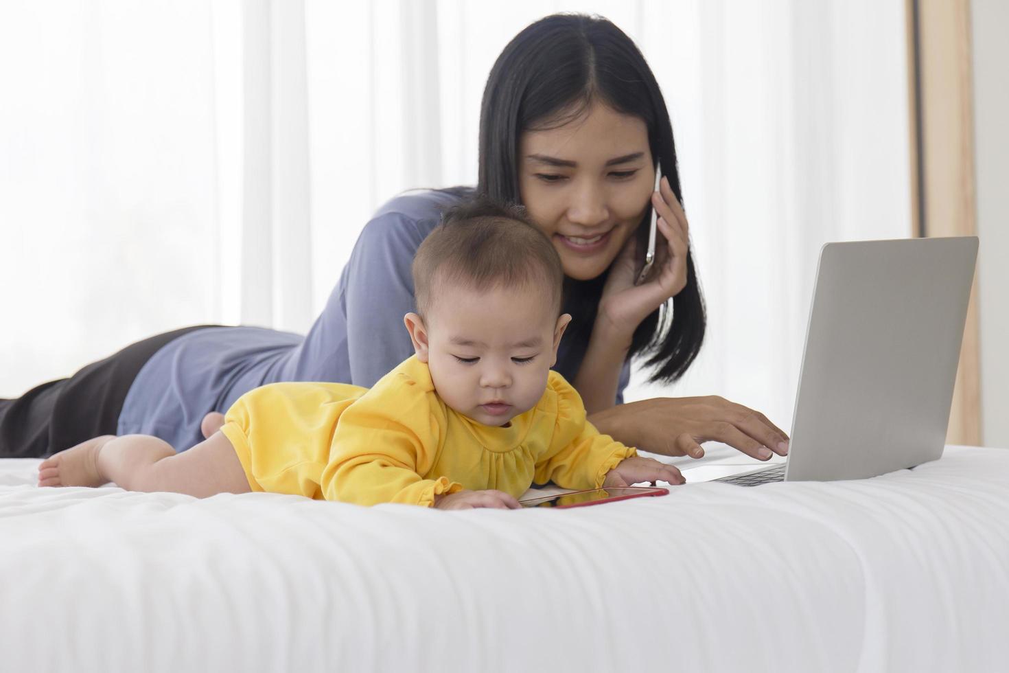 en asiatisk bebis leker bredvid sin mamma i sängen, med sin mamma som pratar i mobiltelefon. foto