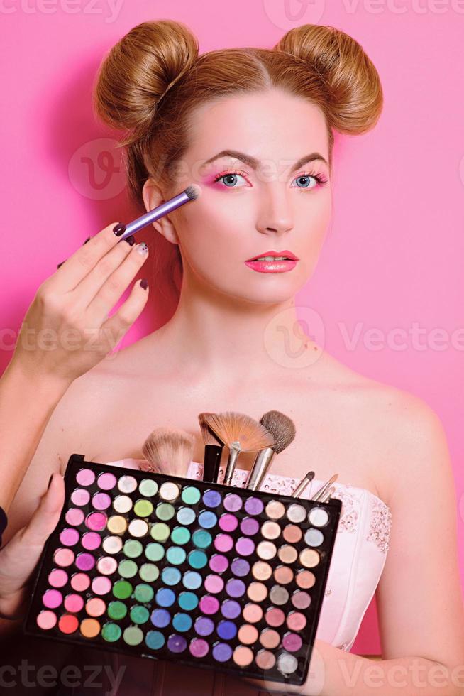 attraktiv blond kvinna med färgglad smink med kosmetiska penslar och skuggor i henne och sminkmästarens händer foto