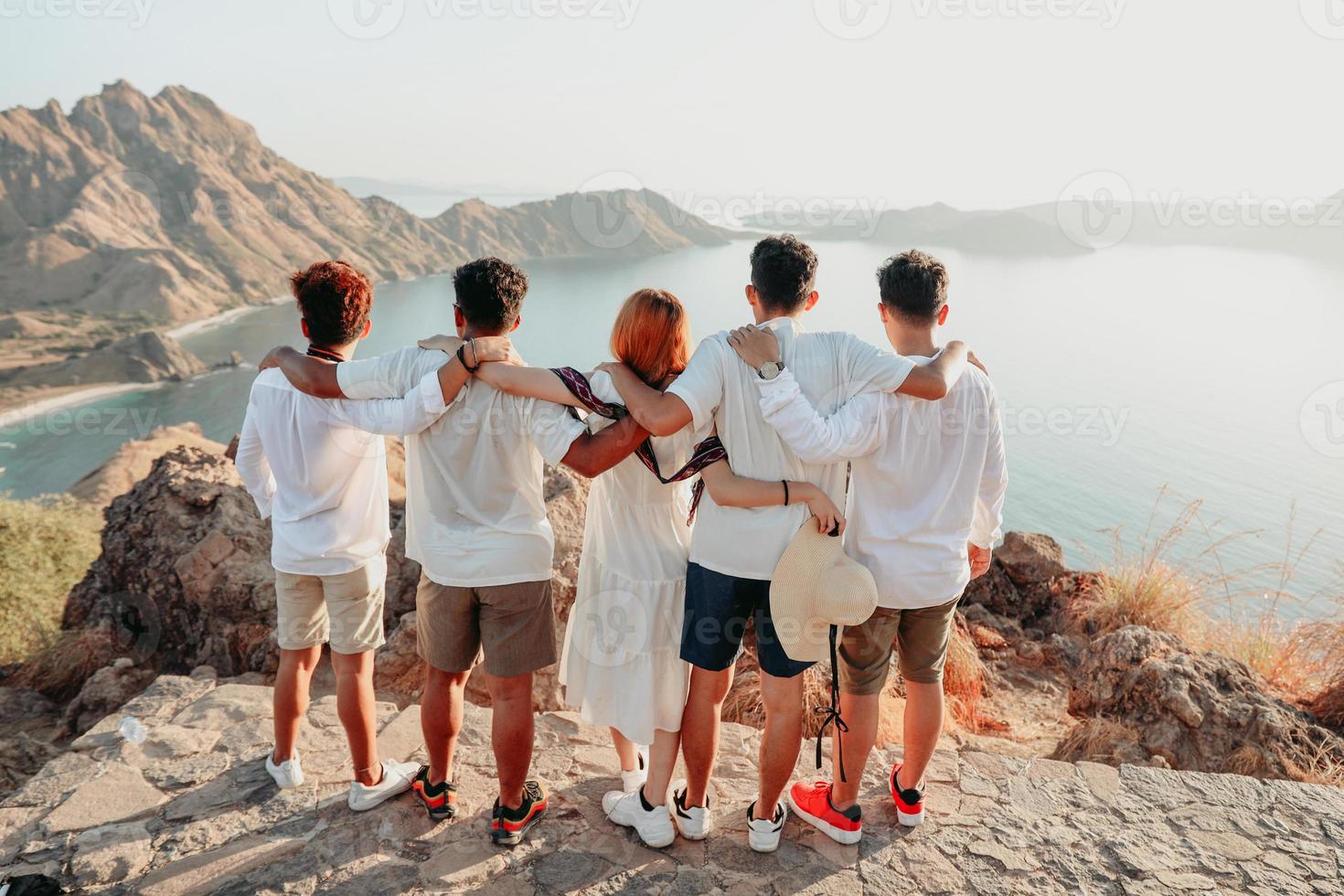 grupp vänner i vit skjorta och klänning som omfamnar varandra på toppen av kullen medan du tittar på landskapet foto