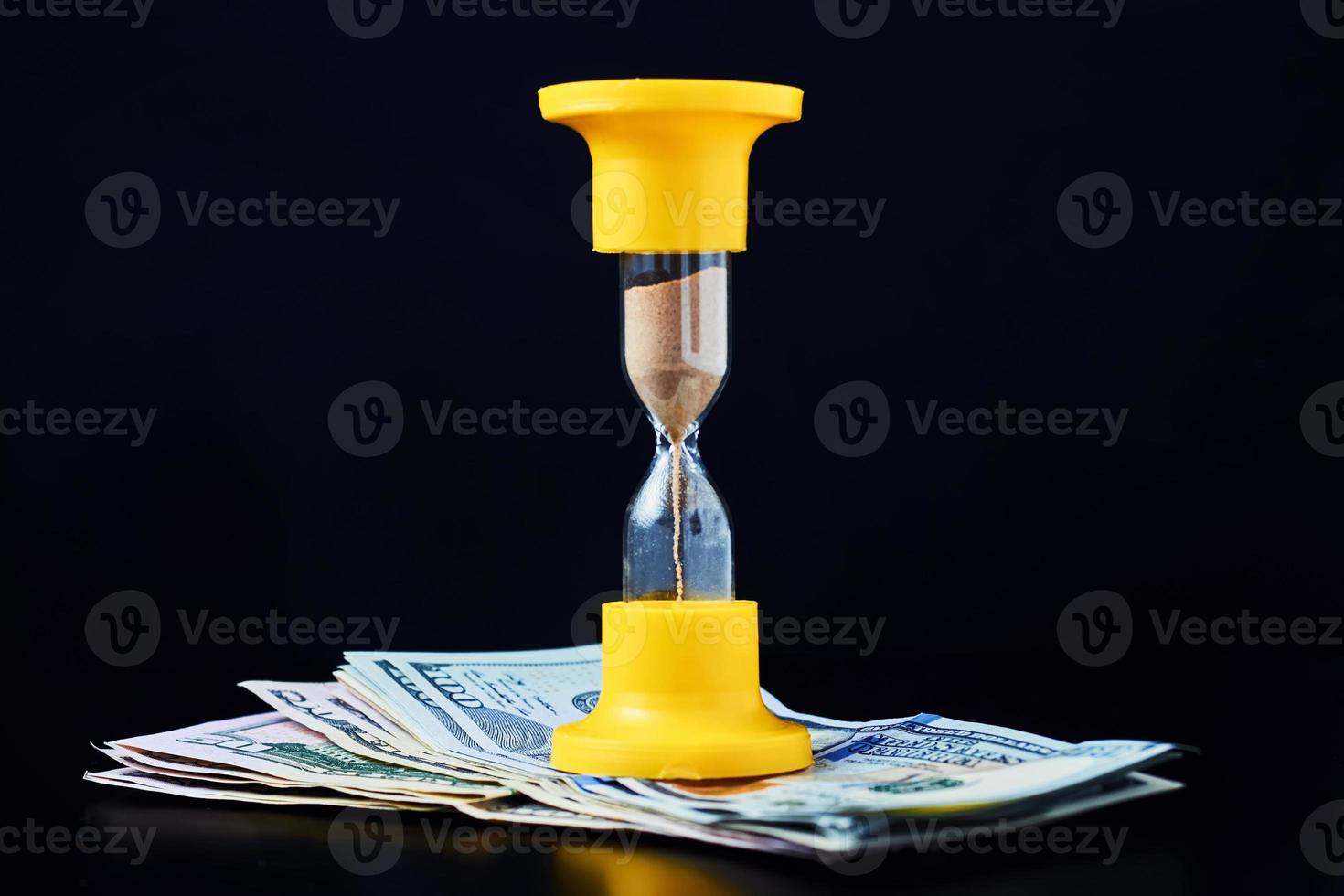 tid är pengar eller tidsinvestering och pensionssparande koncept. gult timglas på högen av dollarsedlar på en mörk bakgrund foto