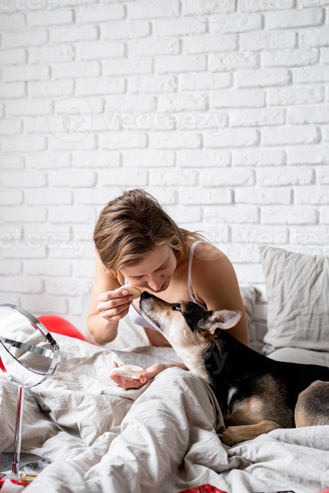 kvinna som sitter på sängen med sin roliga hund hemma och applicerar kosmetika foto