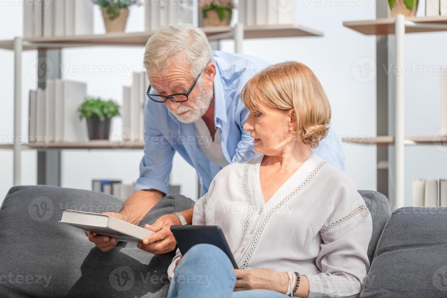 porträtt av lyckliga äldre par i vardagsrummet, mogen kvinna och en man med en bok och surfplatta på mysig soffa hemma, lyckliga familjekoncept foto