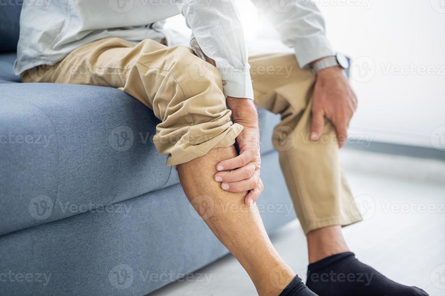 gammal man som lider av knäsmärta sitter soffa i vardagsrummet, äldre man lider av knäsmärta när han sitter i soffan foto