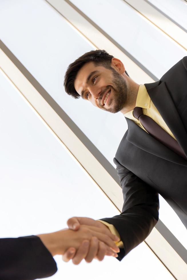 två unga affärsmän är prydligt klädda i kostym och slips, håller varandra i hand och visar separata företag med glada leende ansikten. foto