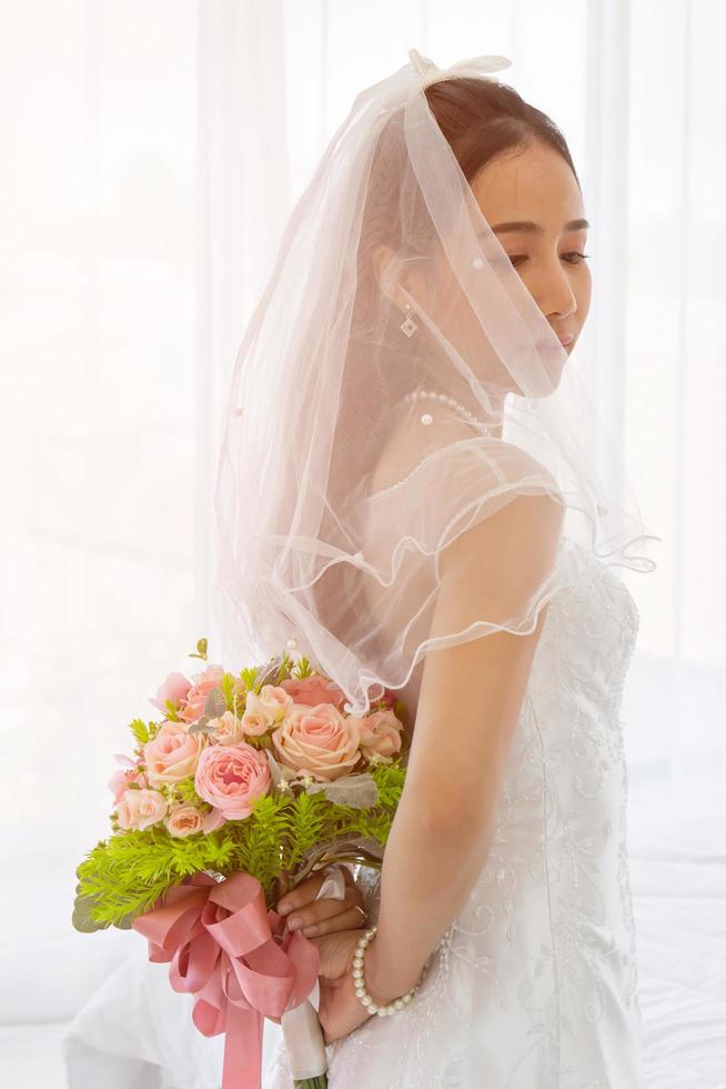 en asiatisk brud bar en vit brudklänning, stående och höll en bukett vackra blommor bakom sig. foto