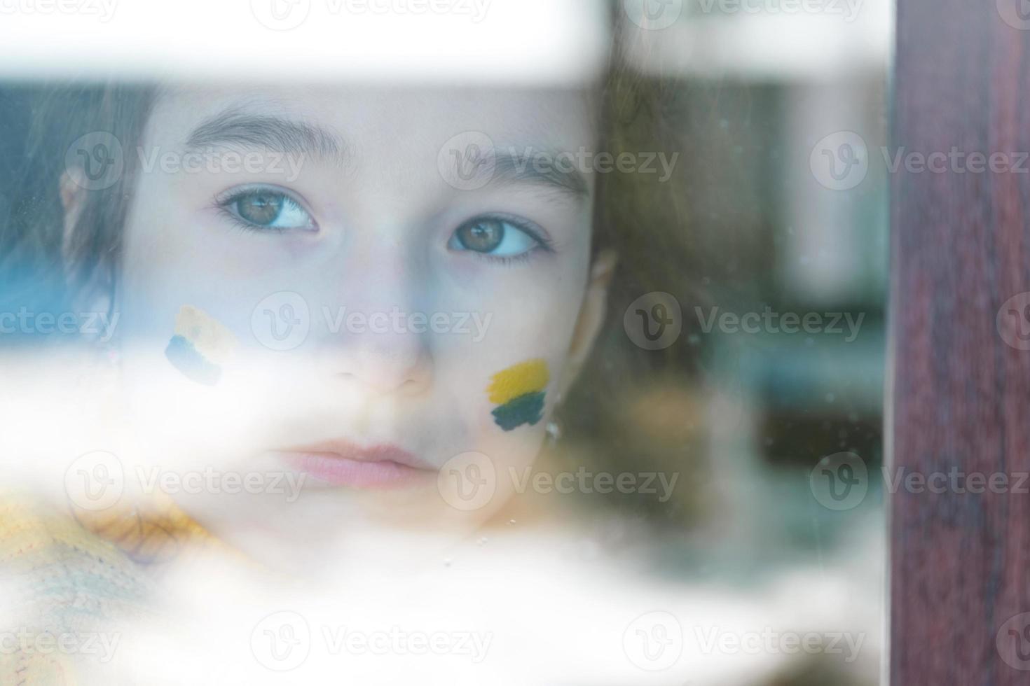 ett ledset barn tittar på fönstret med ukrainska flaggan målad på kinden, oro och rädsla. humanitärt bistånd till barn, världsfred, säkerhet. foto