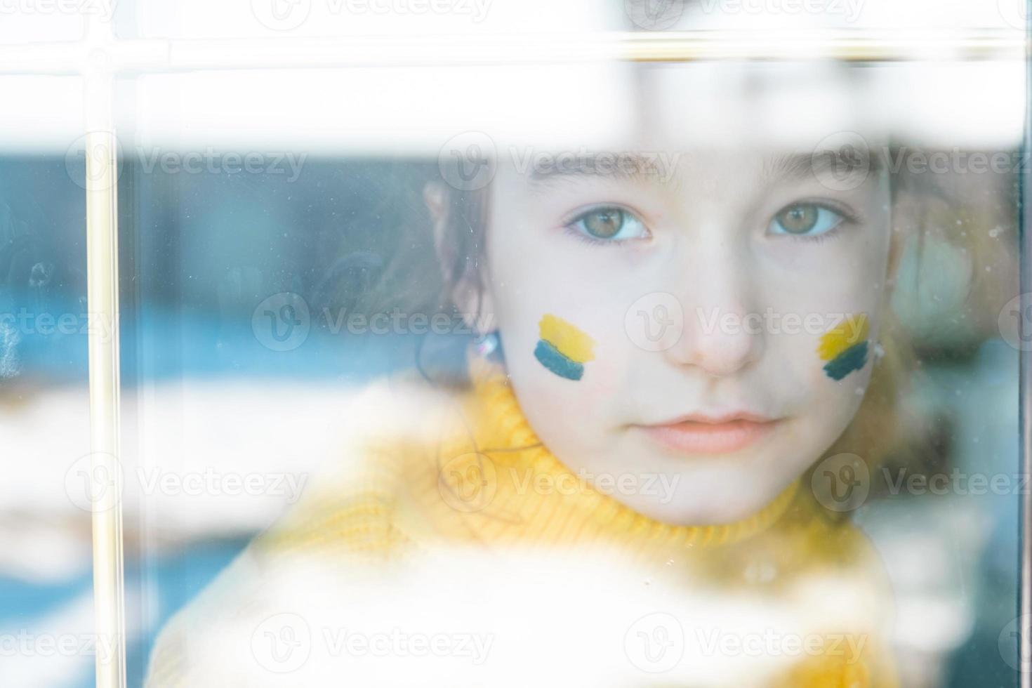 ett ledset barn tittar på fönstret med ukrainska flaggan målad på kinden, oro och rädsla. humanitärt bistånd till barn, världsfred, säkerhet. foto