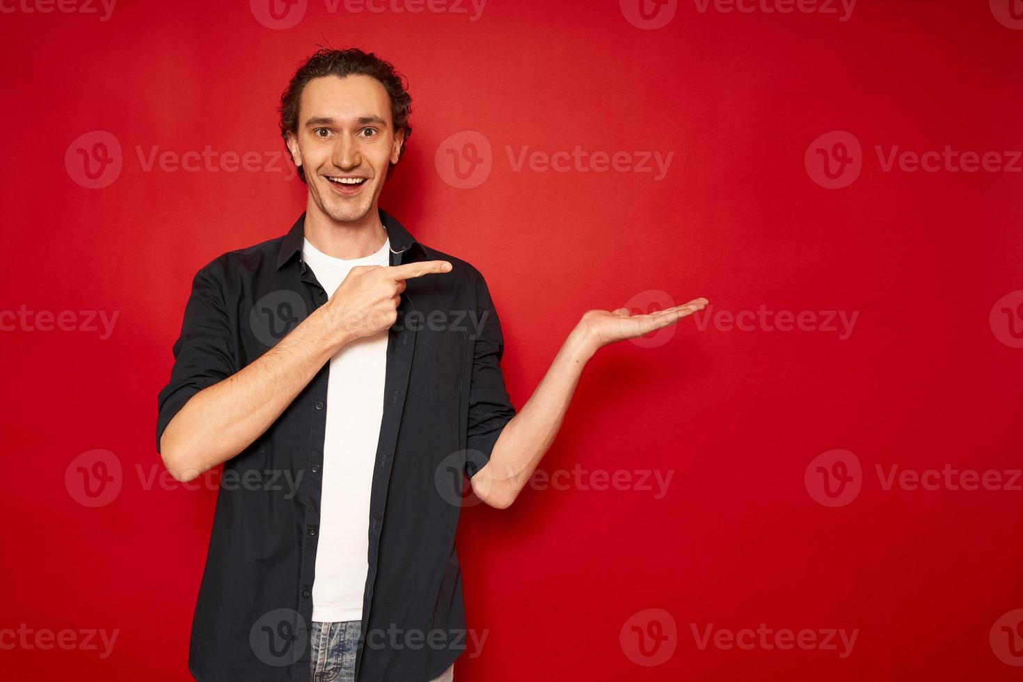 ung, stilig, upphetsad man i vardagskläder, blå skjorta, vit t-shirt, pekar pekfingret mot handfritt tomt utrymme för reklamtextobjekt isolerad röd bakgrund kopia utrymme. rea, promo foto