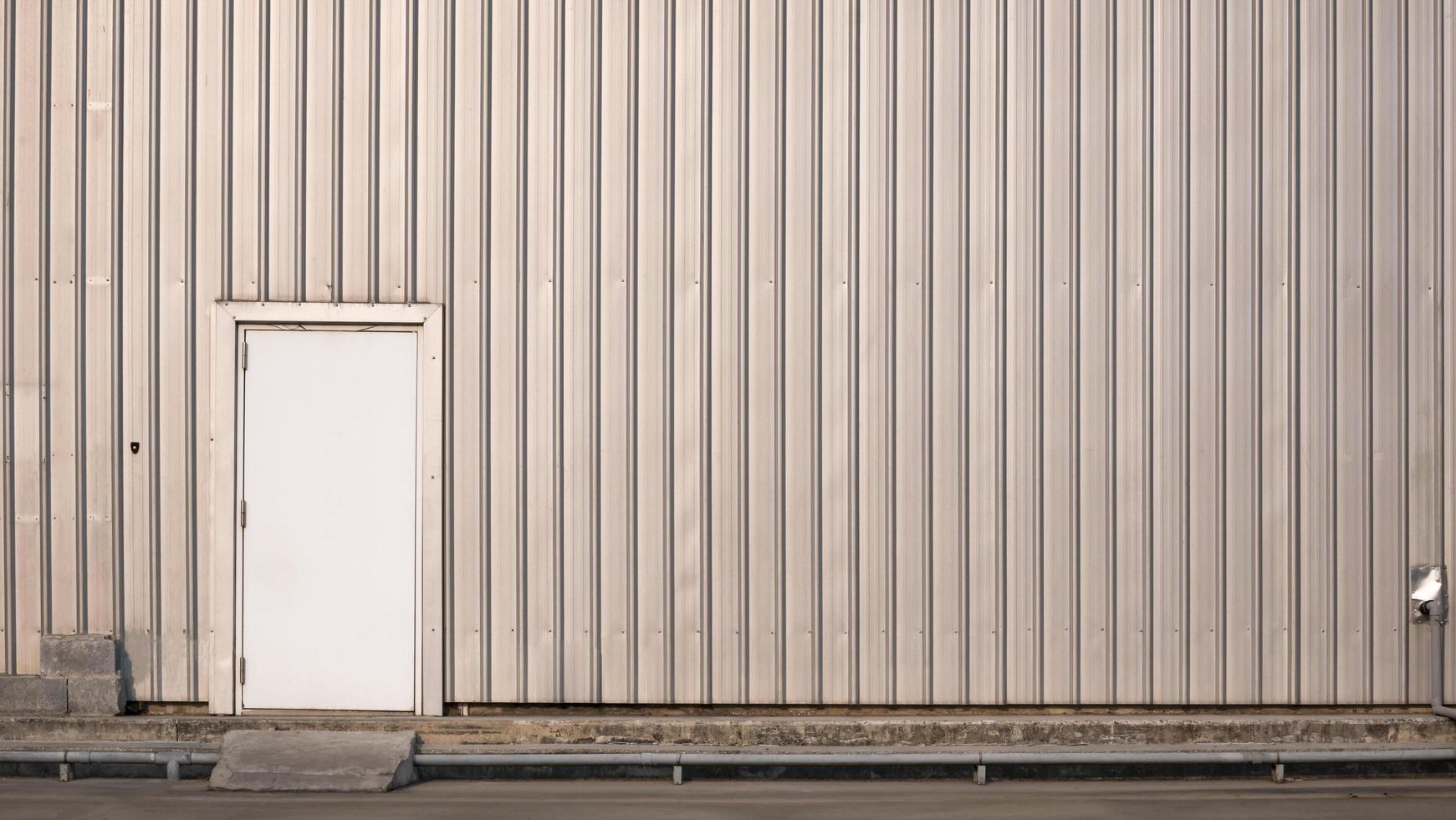 den gamla vita dörren i plåtvägg av lager eller fabriksbyggnad med rörledning på betonggolv foto