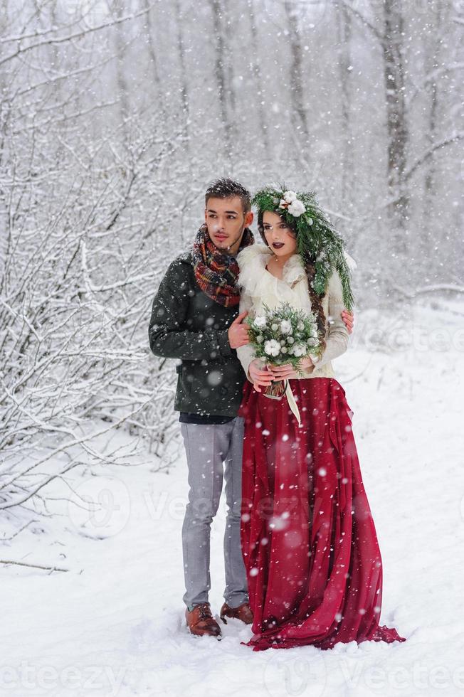 vackra brud och brudgum med en vit hund står på bakgrunden av en snöig skog. foto