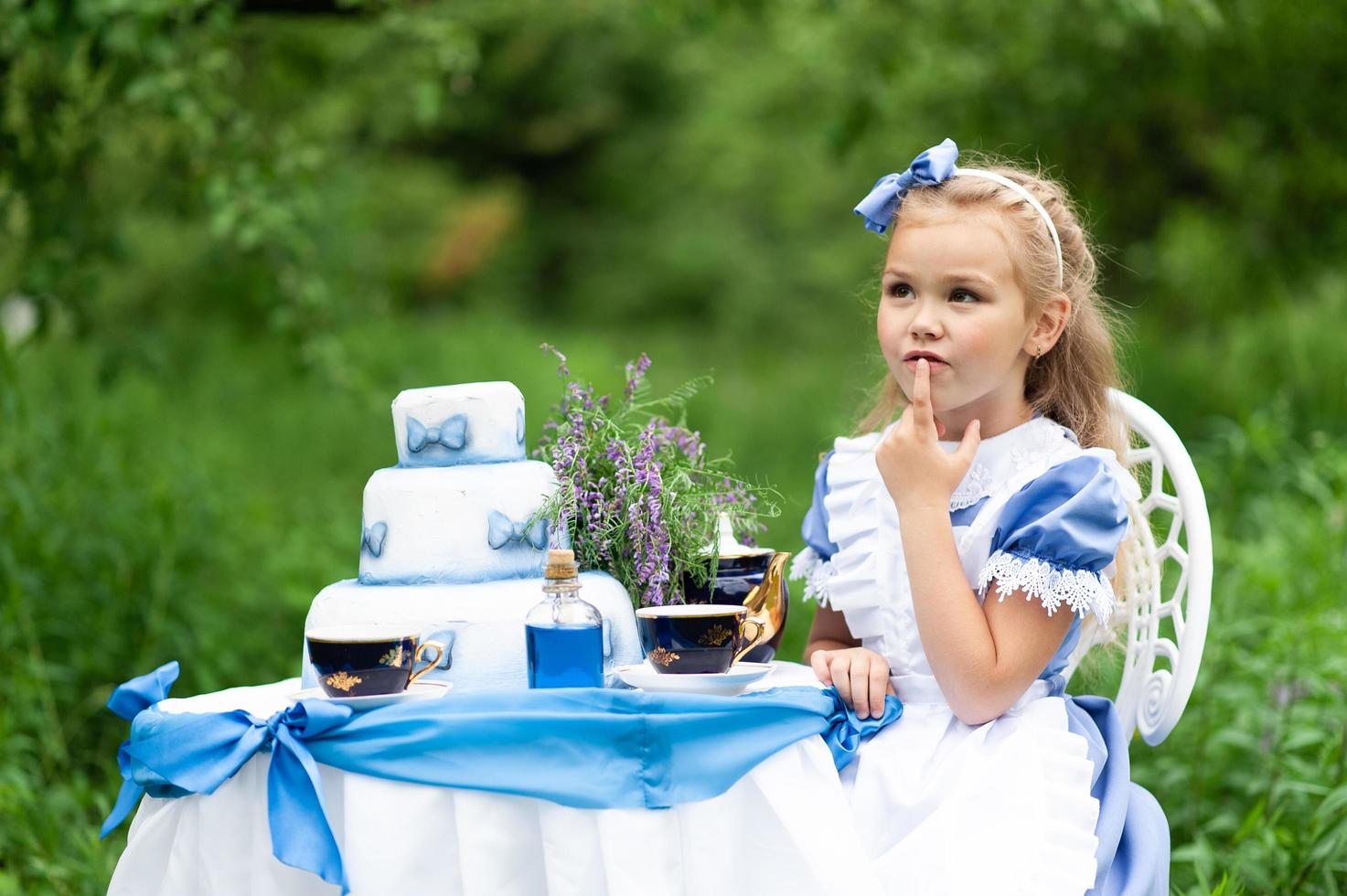 en liten söt tjej i kostymen alice från underlandet håller en tebjudning vid sitt magiska bord. fotograferad i naturen. foto
