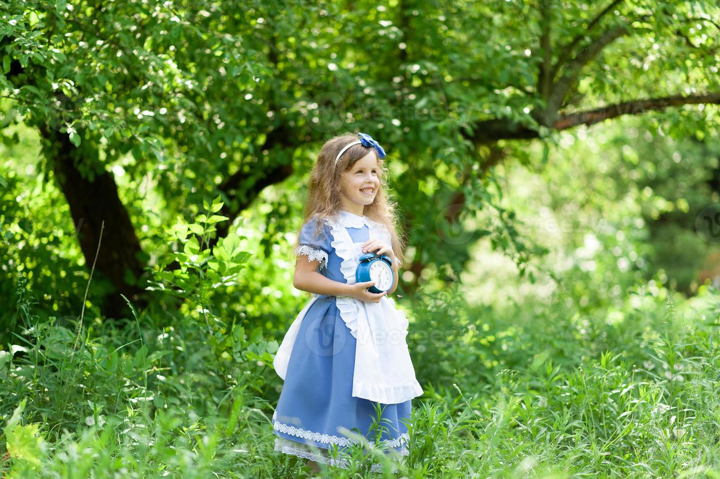 liten söt flicka i en alice kostym håller en gammal antik klocka. foto