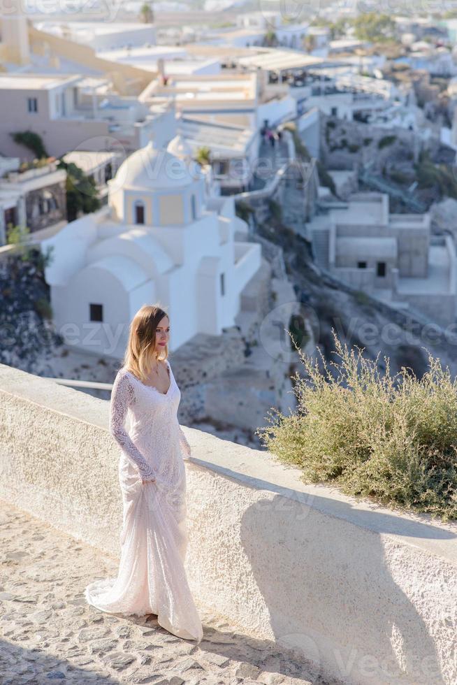 vacker brud i en vit klänning poserar mot bakgrund av staden thira, santorini. foto