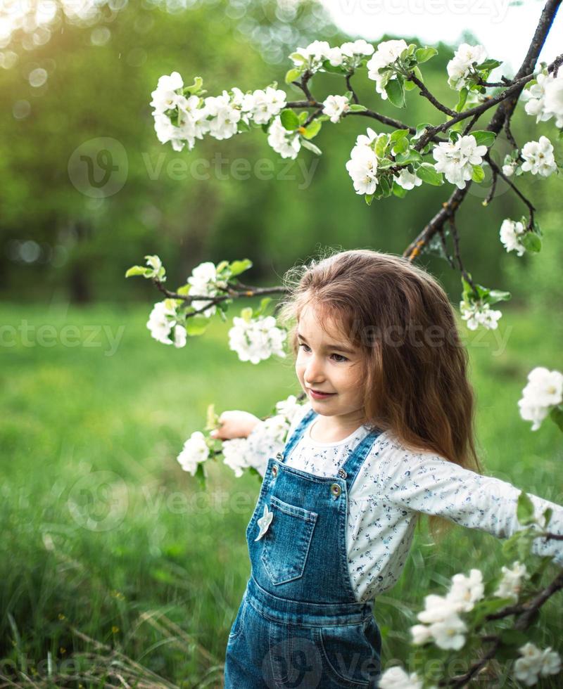 en söt liten flicka på 5 år i en blommande vit äppelodling på våren. vår, fruktträdgård, blomning, allergi, vårdoft, ömhet, omsorg om naturen. porträtt foto