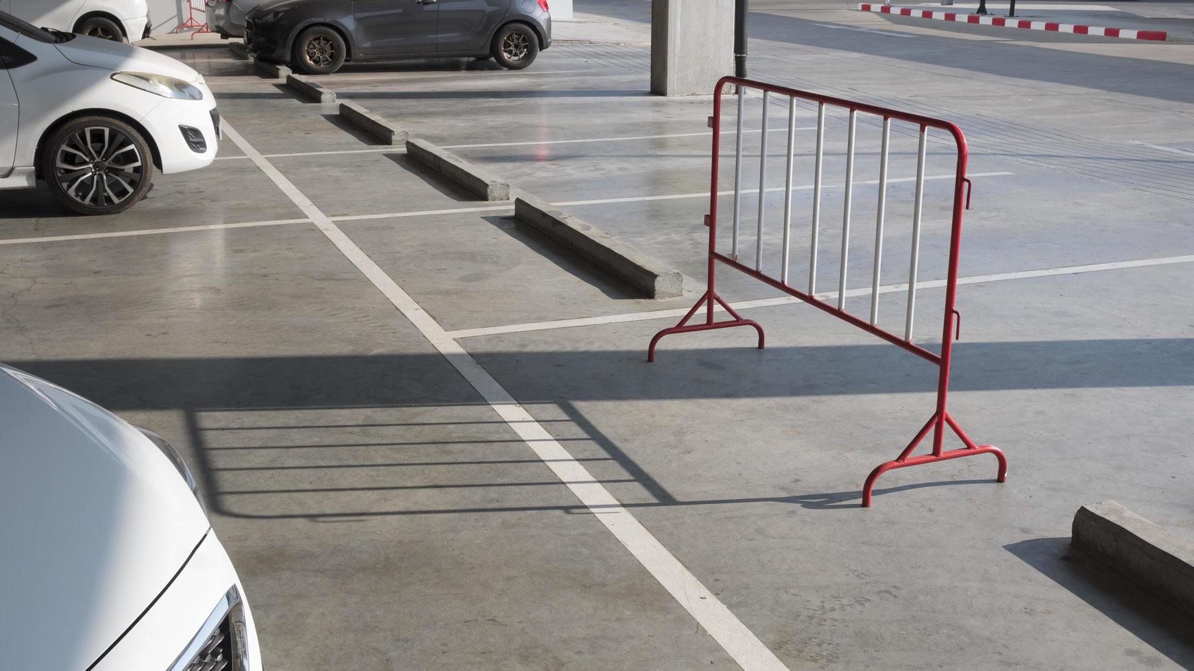 solljus och skugga på ytan av stål röd och vit trafikbarriär med rad av betonghjulstopp och vita markeringslinjer på cementgolv i parkeringsområdet foto