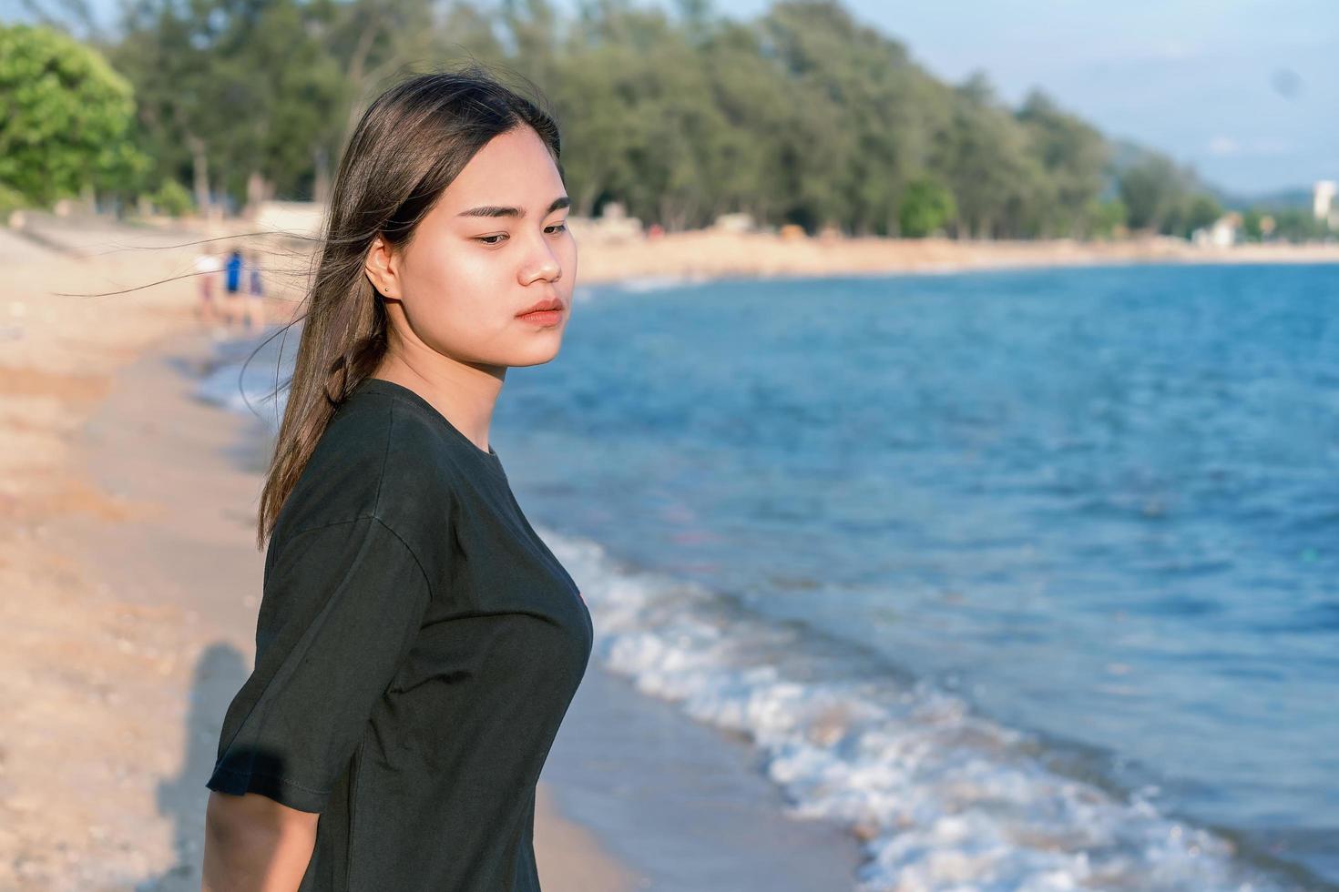 porträtt av en orolig tjej som står på stranden med grön natur och blått hav i bakgrunden. ledsen och deprimerad ung kvinna som ensam står på stranden nära havet. depression koncept. foto