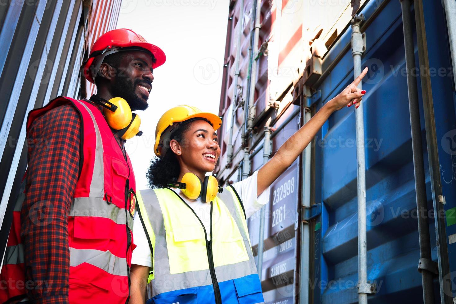 två industriella afroamerikanska ingenjörer man och kvinna bär skyddsväst och hjälm som arbetar tillsammans på logistisk sjöfart last container varv, arbetar kvinnliga pekar och frågar åsikt av kollega. foto